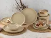 A collection of beige ceramic plates and bowls, some containing potatoes, arranged on a textured surface with dried plants in the background. Produktbild-Vorschau