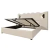 A beige upholstered bed frame with the storage lift mechanism open, revealing the slatted mattress base and storage compartment underneath. Two wall-mounted reading lamps are attached to the headboard. Produktbild-Vorschau