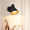 A black wall lamp with a gold interior is mounted on a white wall above some decorative wooden birds and picture frames. Náhľad obrázku produktu