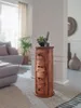 A wooden cylindrical cabinet with drawers stands on a rug in a living room, next to a sofa and a potted plant. Voir les images du produit