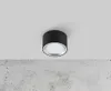 A black cylinder-shaped ceiling light with a silver rim and a white light source is mounted on a white ceiling above a textured concrete surface. Produktbild-Vorschau