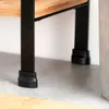 Close-up of the legs and bottom shelf of a wooden shelf unit with black metal legs and black plastic feet, resting on a grey floor. Náhled obrázku produktu