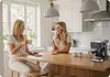 Two women are sitting at a kitchen counter, drinking coffee and chatting. A coffee machine is on the counter. Produktbild-Vorschau