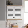 A white cabinet with slatted doors and a wood-tone top is mounted on a tiled wall above laundry detergent bottles and a basket of towels. Náhled obrázku produktu
