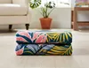 Two folded blankets with a tropical leaf pattern are stacked on a wooden floor in a living room setting. A cream-colored armchair and a potted plant are visible in the background. Podgląd zdjęcia produktu