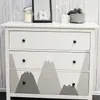 A white dresser with a mountain design on the drawers, decorated with a framed picture of a bear, a candle in a jar, and a small basket with items on top. Náhled obrázku produktu