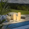 Three LED candles of varying heights are placed on a wooden deck next to a swimming pool, with pillows and a woven pouf nearby. A lush green forest and sky are visible in the background. Podgląd zdjęcia produktu