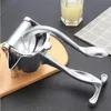 A manual aluminum juicer is placed on a wooden board, with a glass of juice and a napkin in the background. Produktbild-Vorschau