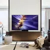 A large television is displayed in a modern living room setting, showcasing its screen with a vibrant abstract image. The TV is placed on a sleek stand, with furniture and decor elements suggesting a home environment. Produktbild-Vorschau