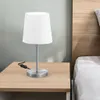 A white fabric lampshade table lamp with a silver base sits on a wooden nightstand next to a bed. Podgląd zdjęcia produktu