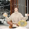 A woman sits in the snow, holding a teapot and a mug, with a cozy sweater and leggings. Snow is visible on the ground and on a window in the background. Produktbild-Vorschau