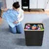 A woman is kneeling on a rug, holding a blue shirt on a hanger. Next to her is a black laundry basket with three compartments filled with clothes. Voir les images du produit