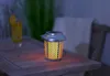 A solar-powered lantern with a flame effect sits on a wooden table next to a drink. Produktbild-Vorschau