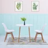 A white round dining table with two white chairs and a vase with green plants on it, set against a light blue paneled wall and a pink upper wall. Náhled obrázku produktu