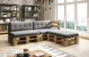 A sectional sofa made from wooden pallets with gray cushions, placed in a living room with a brick wall and large windows. Produktbild-Vorschau