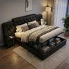 A bedroom scene with a large, tufted black bed as the focal point, featuring storage underneath and bedside tables with lamps. Produktbild-Vorschau
