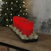 Four red LED candles are arranged on a black tray with moss, placed on a wooden surface in front of a Christmas tree with lights. Produktbild-Vorschau