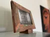 A wooden picture frame with a black and white photo inside, resting on a glass surface with a painting in the background. Produktbild-Vorschau