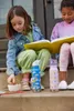 Two young girls are sitting on steps, reading a book and eating snacks. Two water bottles are placed in front of them. Voir les images du produit