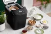 A black Panasonic bread maker is shown on a kitchen counter, surrounded by ingredients for baking bread. Náhľad obrázku produktu