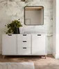 A white sideboard with drawers and doors is placed against a wall with a floral wallpaper. A black framed mirror hangs above the sideboard, reflecting a wooden beam. A potted plant and books are on top of the sideboard. Produktbild-Vorschau