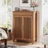 A wooden shoe cabinet with slatted doors and a drawer is placed in a bedroom setting next to a bed and a window. Náhľad obrázku produktu