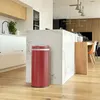 A modern kitchen with a red automatic trash can in the foreground, placed next to a marble island. The background shows kitchen cabinets, a sink, and a dining area with chairs. Produktbild-Vorschau