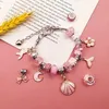 A collection of pink and silver charms and beads for making bracelets and necklaces, laid out on a pink surface with a piece of paper and wheat stalk in the background. Produktbild-Vorschau
