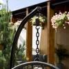 A close-up of a black metal chain and spring attached to a black curved stand, part of a hanging chair setup. In the background, there are green trees and hanging pink flowers. Náhled obrázku produktu