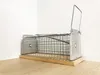 A metal live trap for rodents sits on a wooden surface against a white wall. Produktbild-Vorschau
