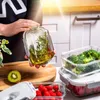A person is holding a glass container filled with olive oil and herbs, with other glass containers filled with broccoli and raspberries in the background. Anteprima dell’immagine prodotto