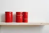 Three red ceramic containers with wooden lids are displayed on a light-colored shelf against a white background. Produktbild-Vorschau