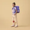 A young girl wearing a backpack stands against a plain yellow background. Produktbild-Vorschau