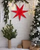 A red paper star decoration hangs on a white brick wall, with Christmas trees and gifts in the foreground. Produktbild-Vorschau