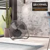 A black floor fan is placed on a rug in a room with a brick wall and a large potted cactus. Produktbild-Vorschau
