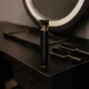 A black hair trimmer with gold accents sits on a black surface, with two brushes and a blurred mirror in the background. Náhled obrázku produktu