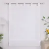A white sheer curtain hanging on a curtain rod against a white wall, with a lamp and plants in the background. Produktbild-Vorschau