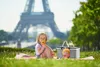 A young girl is having a picnic with a doll and a picnic basket in front of the Eiffel Tower. Produktbild-Vorschau