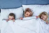 A mother and her two children are peeking out from under a white duvet on a bed. Produktbild-Vorschau