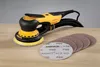 A yellow and black orbital sander with a black hose is placed on a wooden surface next to several circular sanding discs. Produktbild-Vorschau