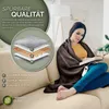 A woman is sitting on a couch wrapped in a brown heated blanket, reading a book. An infographic on the left shows the layers of the blanket. Produktbild-Vorschau
