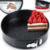 A close-up of a black springform baking pan with a slice of cake topped with strawberries inside it. There is also a smaller springform pan with jelly on top in the background, and packaging for the baking tin in the top left corner. Náhled obrázku produktu