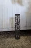 A black solar-powered LED pathway light with a decorative cut-out pattern stands on a gravel surface in front of a white corrugated wall. Produktbild-Vorschau