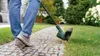 A person is using a grass trimmer along a stone pathway in a garden. Produktbild-Vorschau