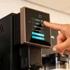 A close-up of a person's finger interacting with the touchscreen display of a coffee machine. Produktbild-Vorschau