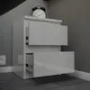 A white bedside table with two drawers, one partially open, sits against a gray wall. An alarm clock is on top of the table. Voir les images du produit