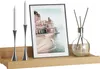 A wooden shelf with decorative items including two candlesticks with candles, a framed picture of a coastal town, and a diffuser with reeds. Produktbild-Vorschau