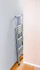 A folded aluminum step stool is placed in the corner of a room, leaning against a light gray wall. A light pink wall is visible to the right, and a wooden floor is at the bottom. Produktbild-Vorschau