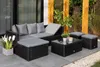 An outdoor patio setting with a black rattan lounge set, including a sectional sofa, coffee table, and two ottomans. The furniture is arranged on a gray tiled patio, with a blurred background of greenery and wooden posts. Náhled obrázku produktu