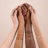 Four hands of different skin tones are held together, with foundation swatches on the forearms. Produktbild-Vorschau